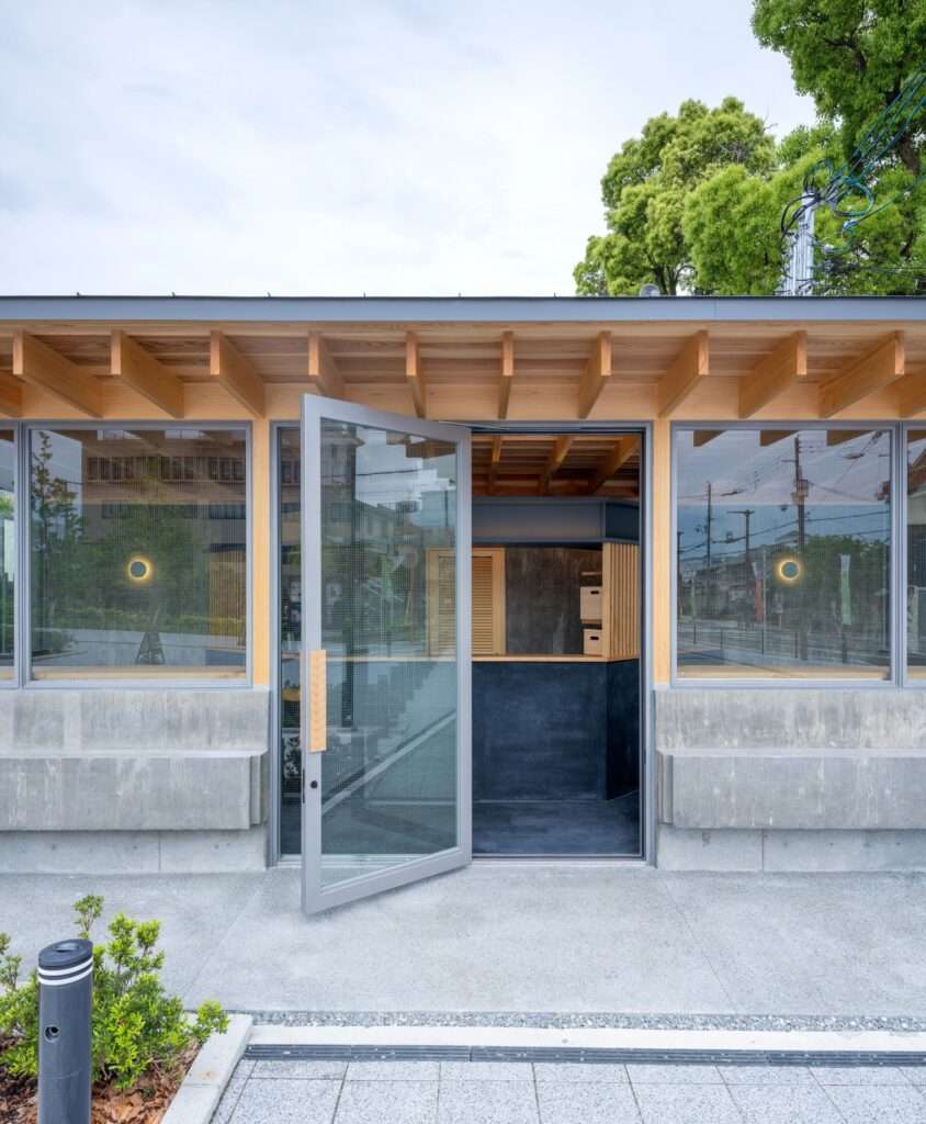 Open glass entrance door of Cafe N+ showcasing timber rafter ceiling details, mesh-reinforced glass windows, and a concrete bench base.