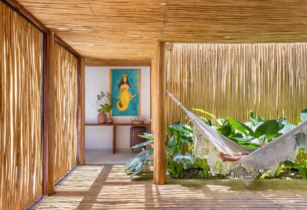 A person relaxing in a white hammock on a wooden deck terrace with biriba wood partition screens and lush tropical foliage.