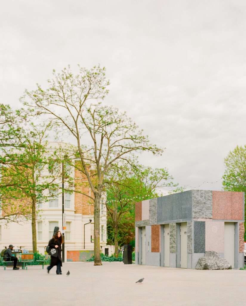 Maida Hill public amenity pavilion clad in multicolored reclaimed stone blocks in a paved public square in London.