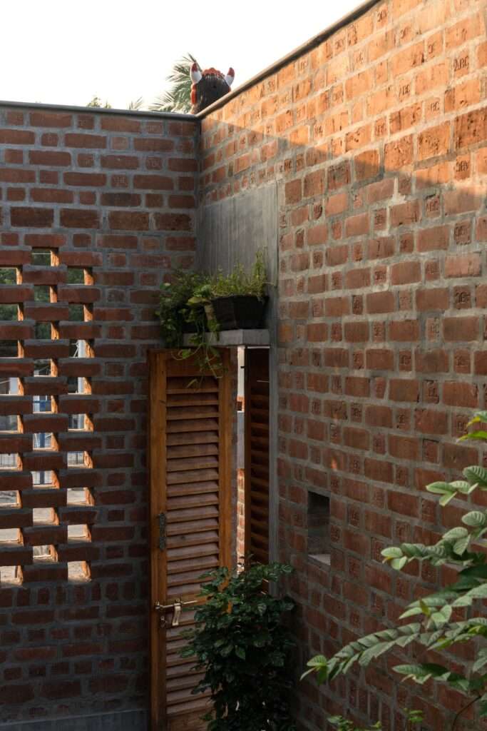 External courtyard corner of Thuraiyur Farmhouse, showing exposed load-bearing brickwork, a perforated brick jali screen, a louvred wooden door, and potted greenery.