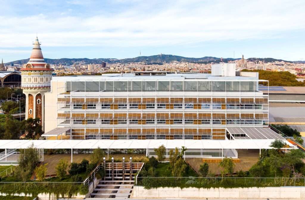 Front elevation of Hospital del Mar expansion showing modular louvered facade, green rooftop garden, and historic water tower in Barcelona.