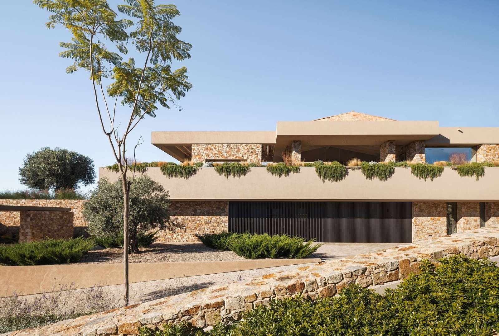 Talleru House modern exterior facade featuring dry-stacked local stone walls, upper sand-toned finish, and landscaped driveway.