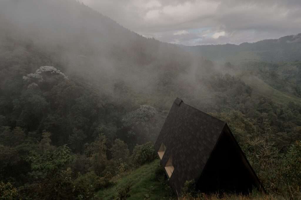 High-angle aerial view of a dark wooden A-frame shelter nestled on a grassy slope within a misty Andean cloud forest in Campano Frío.