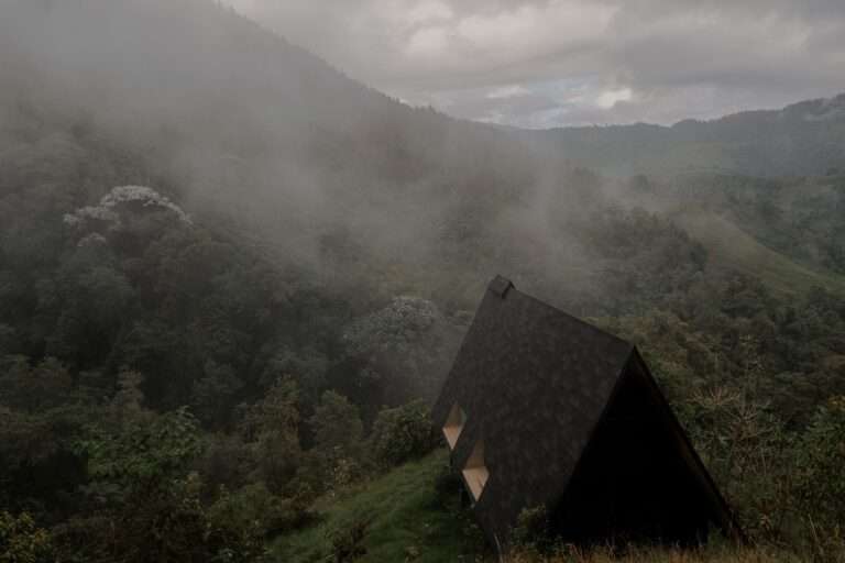 High-angle aerial view of a dark wooden A-frame shelter nestled on a grassy slope within a misty Andean cloud forest in Campano Frío.