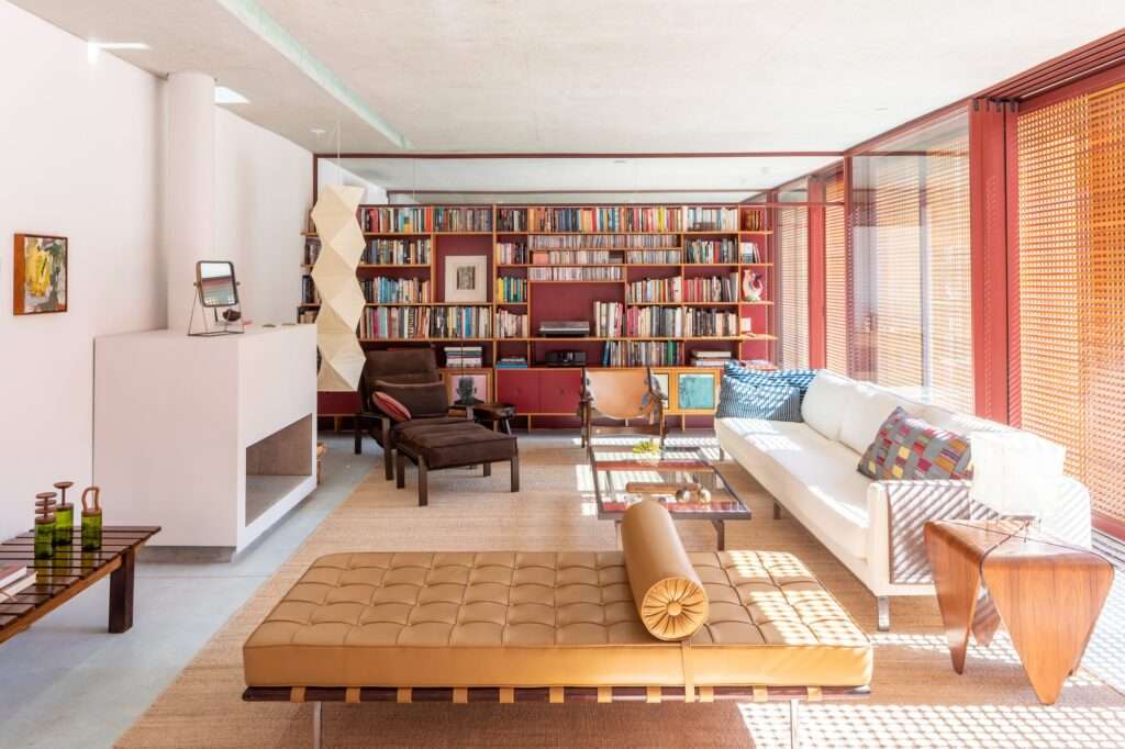 Modern open-plan living room with a white fireplace, floor-to-ceiling bookshelf wall, mid-century furniture, and light filtered through wooden screens.