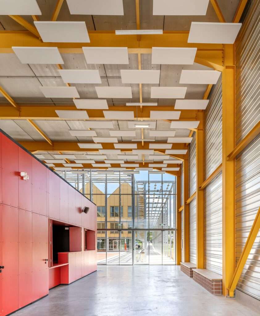 High-ceiling interior hall featuring yellow steel structural beams, suspended acoustic ceiling panels, and a red reception booth looking out onto an exterior plaza.
