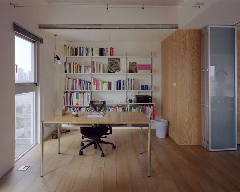 Dedicated home office study space featuring a wooden desk, black ergonomic chair, floor-to-ceiling bookshelf, wood plank flooring, and semi-translucent accordion sliding door.