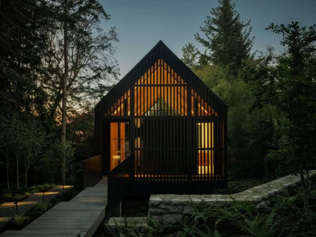 Front view of Forest Studio at dusk showing a dark slatted wooden facade, gabled roof, warm interior light, and an elevated wooden walkway entrance.