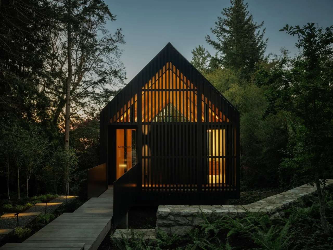 Front view of Forest Studio at dusk showing a dark slatted wooden facade, gabled roof, warm interior light, and an elevated wooden walkway entrance.