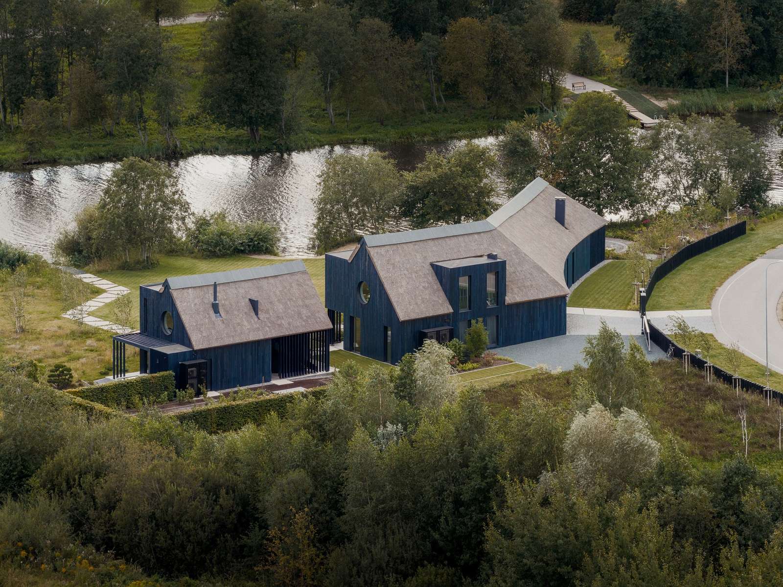 Aerial view of BPROM Residence featuring charrred dark timber cladding, thatched roofs, and curved volumes next to a river.