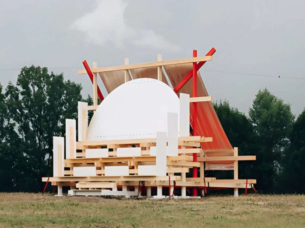 Daytime exterior view of the Gabonsa Pavilion, a temporary wooden structure on a grassy field, featuring a white dome element, red supporting beams, and reflective orange mesh under a cloudy sky.