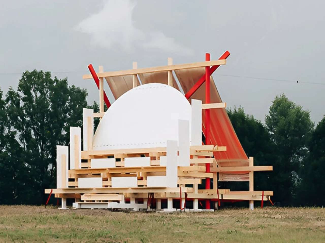 Daytime exterior view of the Gabonsa Pavilion, a temporary wooden structure on a grassy field, featuring a white dome element, red supporting beams, and reflective orange mesh under a cloudy sky.