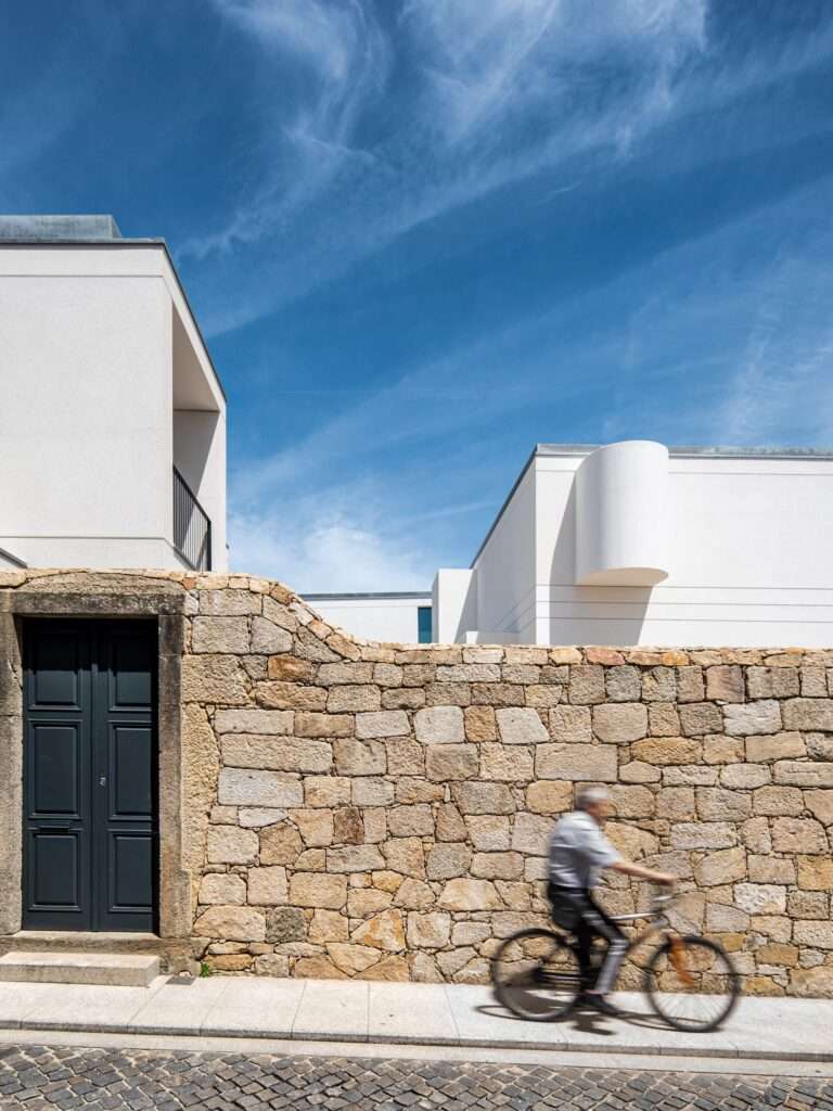 Historic granite wall with a dark wooden doorway in front of a white contemporary house volume in Vila do Conde.