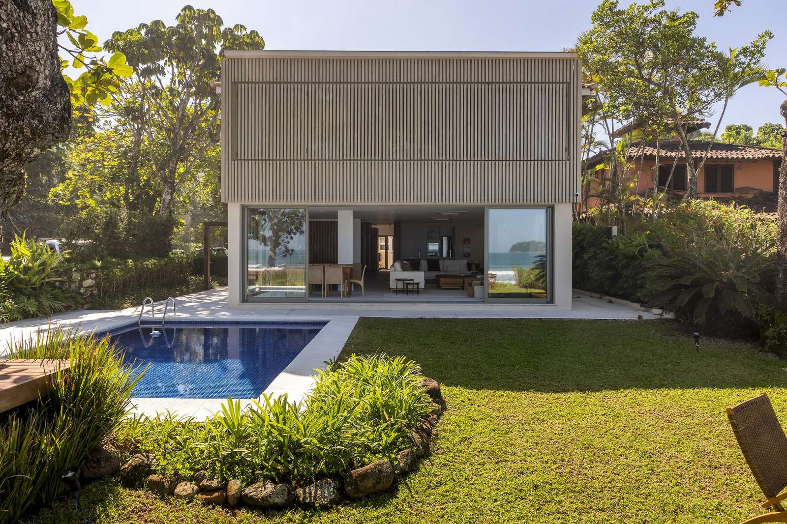 Rear elevation of Casa ADS showing a swimming pool, green lawn, open sliding glass doors on the ground floor, and closed wooden louvered shutters on the upper floor.