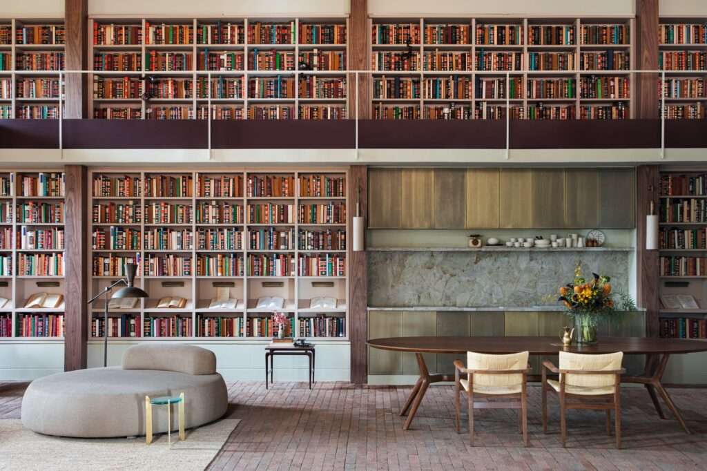 Double-height central library with wall-to-wall wooden bookshelves integrated beside a dining table and kitchen niche in Casa Curral Cileiro Alvorada.