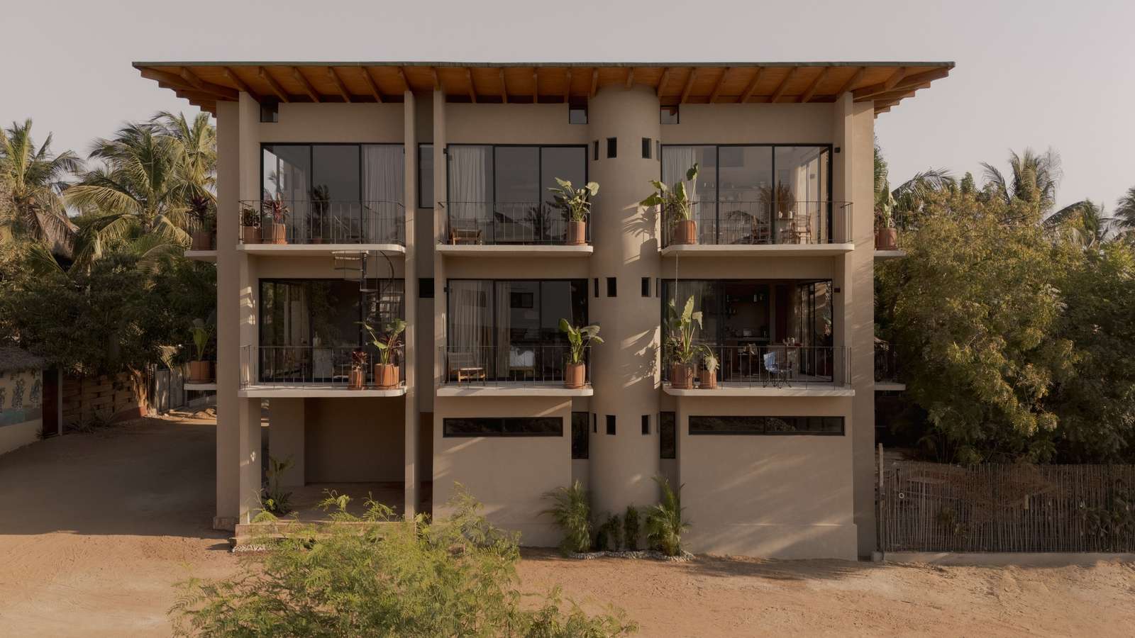 Front facade elevation of Suuel Living building showing multi-level concrete balconies, potted plants, and large sliding glass doors in a coastal landscape.