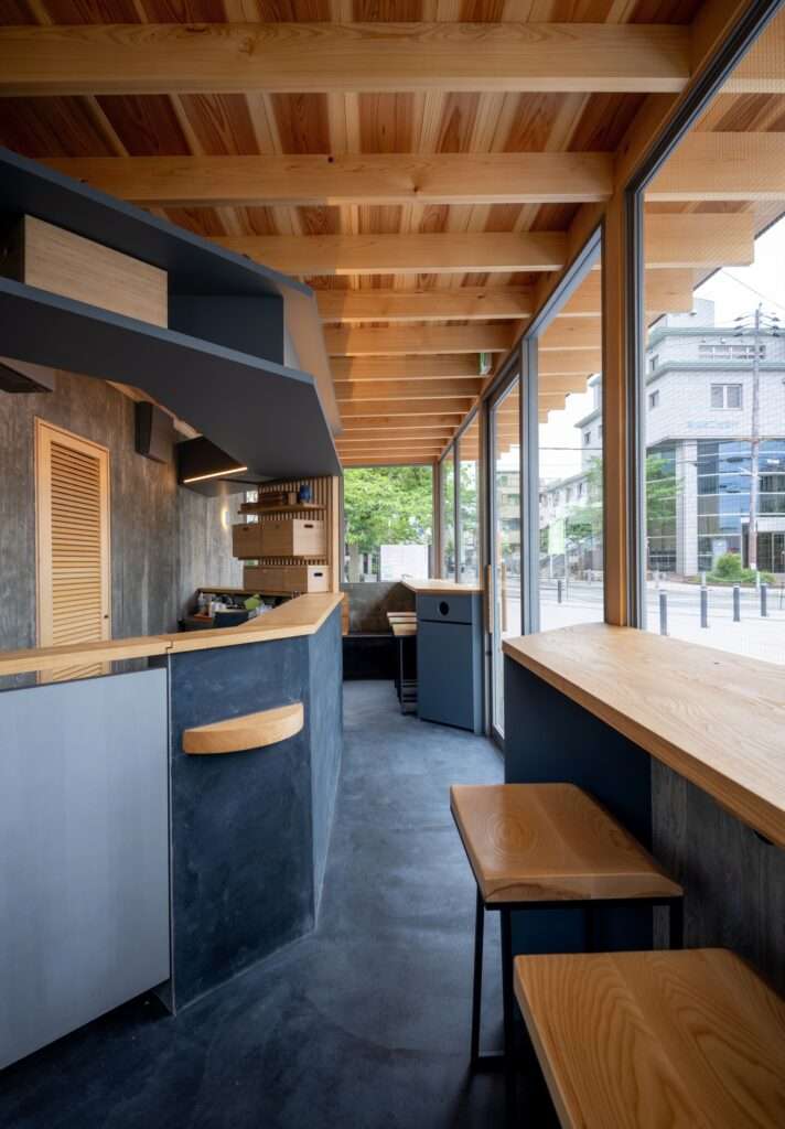 Interior corridor of Cafe N+ featuring dark concrete floors, wooden ceiling rafters, a dark service counter, and wooden bar seating facing the street.