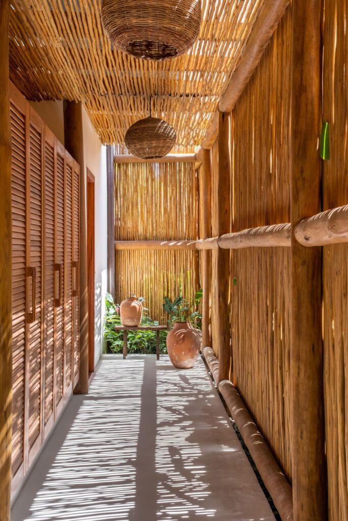 Covered architectural corridor featuring a biriba branch ceiling, slatted wooden wardrobe doors, terracotta pots, and natural light shadow patterns on the floor.