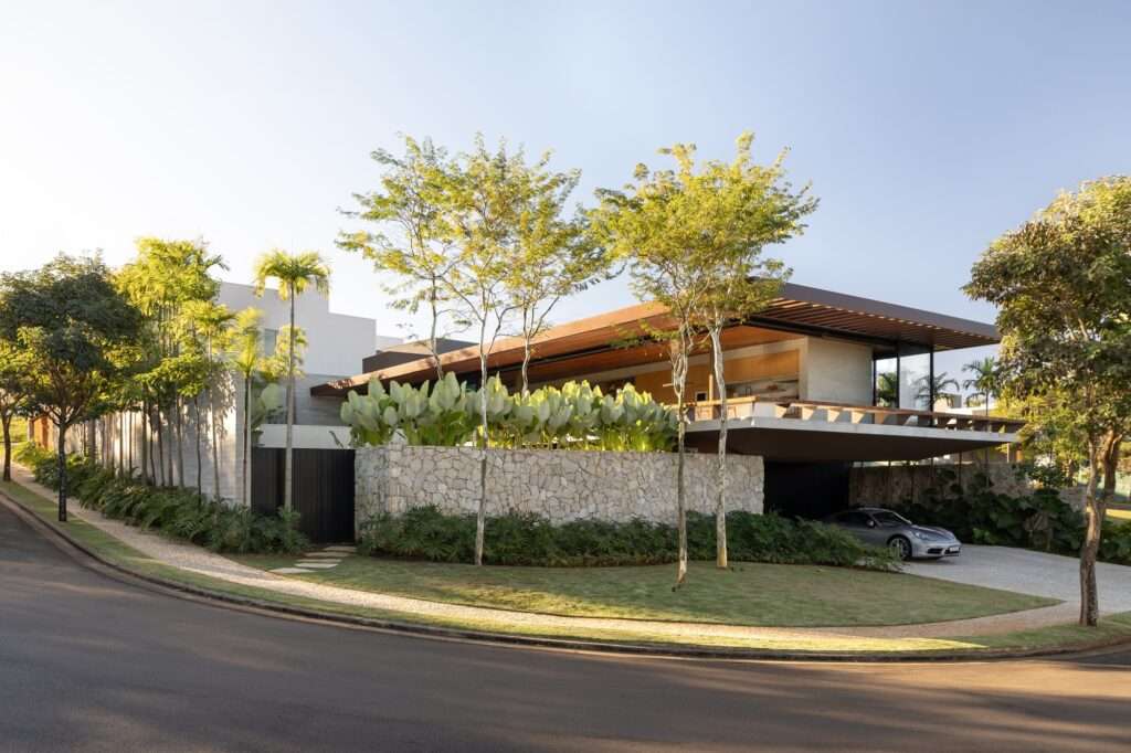 Wide street-level perspective of Casa da Mata positioned on a sloping corner site enclosed by tropical trees and stone boundary walls.