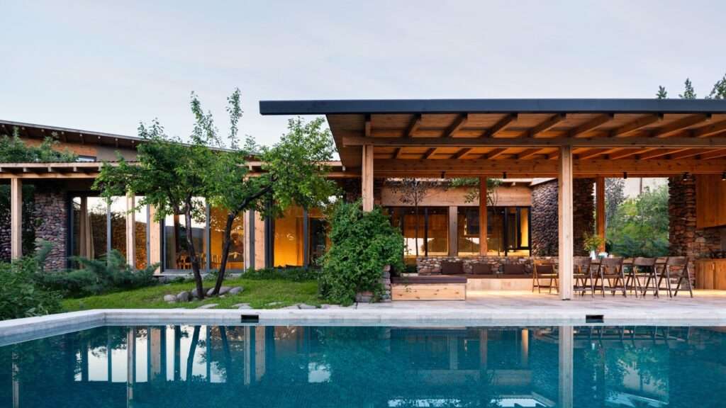 Outdoor swimming pool terrace and shaded wooden pavilion of Buladala House featuring stone walls and natural timber structure.