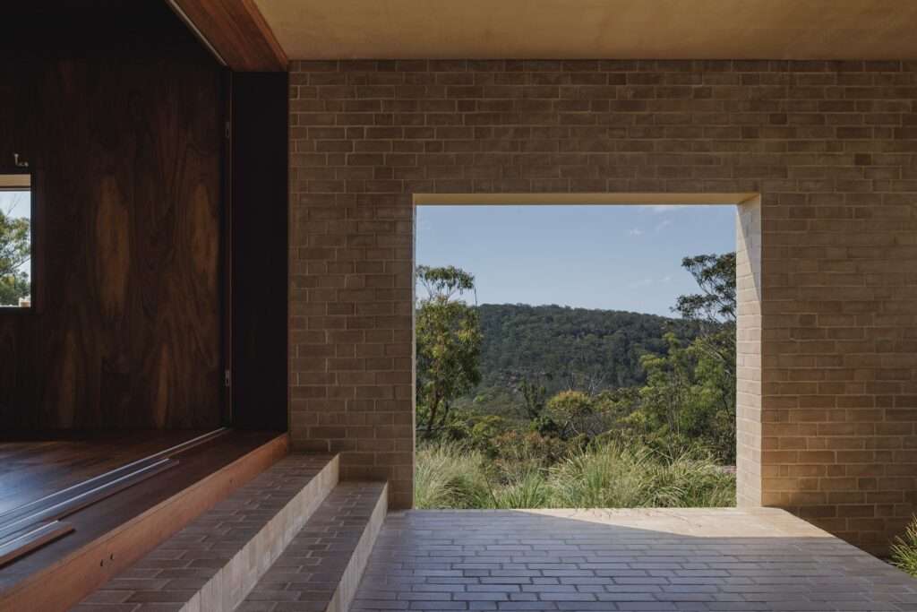 A framed view of a forested hill and blue sky seen through a square opening in EA House's brick-paved external terrace and facade wall.