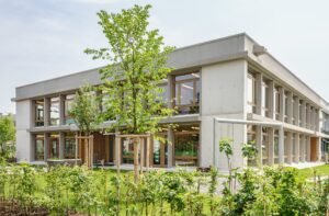 Exterior of the two-story Mösli school extension in Ostermundigen featuring a sandblasted concrete frame, floor-to-ceiling wooden windows, and surrounding green landscaping.