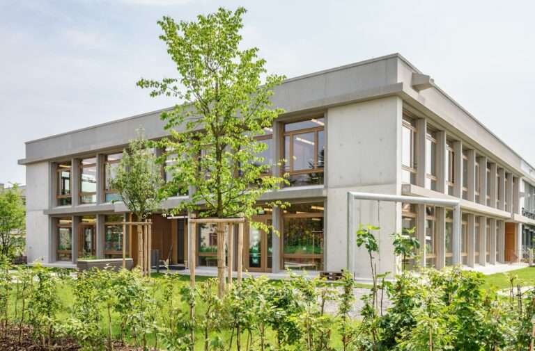 Exterior of the two-story Mösli school extension in Ostermundigen featuring a sandblasted concrete frame, floor-to-ceiling wooden windows, and surrounding green landscaping.