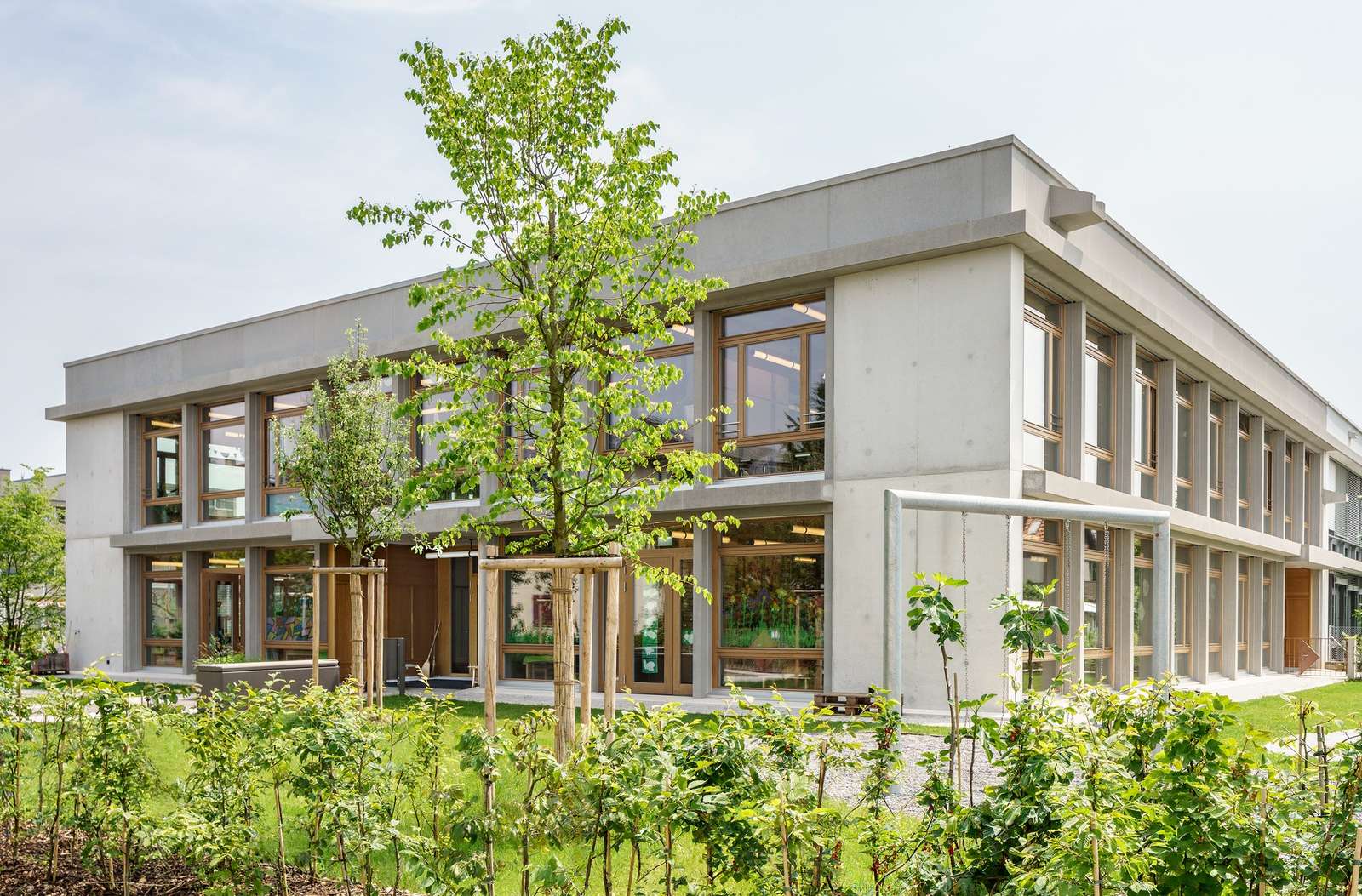 Exterior of the two-story Mösli school extension in Ostermundigen featuring a sandblasted concrete frame, floor-to-ceiling wooden windows, and surrounding green landscaping.