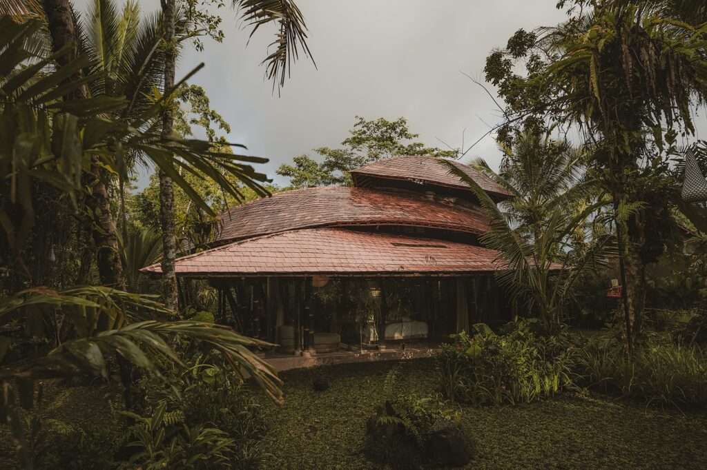 Exterior wide view at dusk of a multi-tiered tile and bamboo roof structure of the Hideout Leaf cabin, surrounded by a lush tropical forest under an overcast sky.