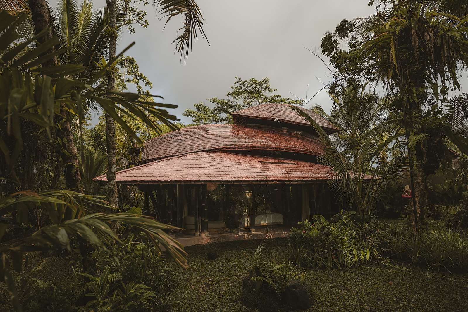 Exterior wide view at dusk of a multi-tiered tile and bamboo roof structure of the Hideout Leaf cabin, surrounded by a lush tropical forest under an overcast sky.