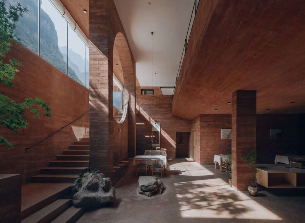 Double-height interior dining hall of Wu Quan Shan Fang featuring red board-formed concrete walls, natural stone boulders, a large circular wall cutout, and high windows framed against mountain cliffs.