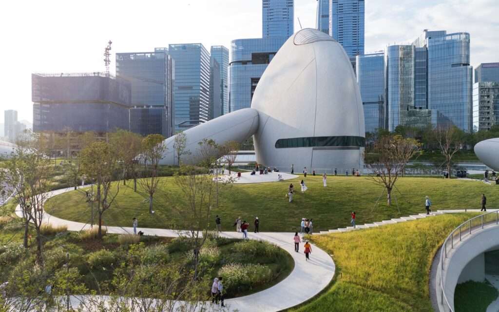 Visitors walking along green sloped lawns and curved pedestrian paths in front of a white egg-shaped pavilion at Shenzhen Bay Culture Square, set against modern city skyscrapers.