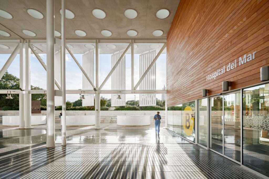 Semi-outdoor entrance plaza of Hospital del Mar Expansion featuring white structural columns, wooden cladding, and skylights.