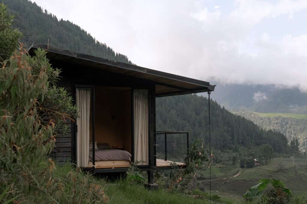 A wooden cabin shelter with wide open sliding glass doors and a bedroom overlooking a dense mountain forest landscape.