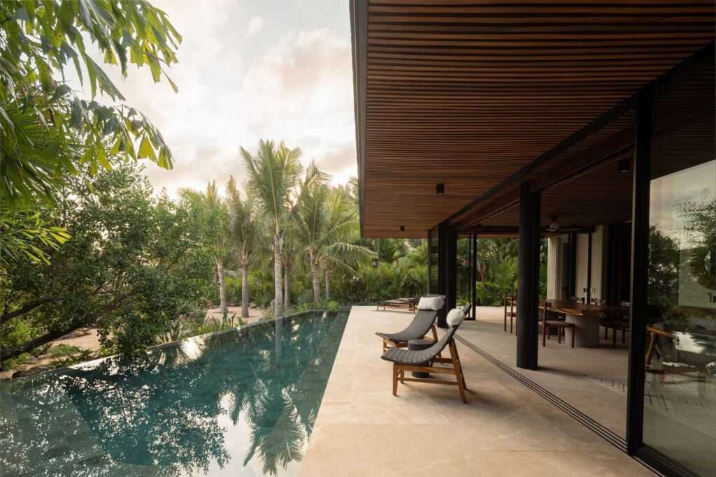 Open outdoor terrace with a wooden slatted ceiling, lounge chairs, and an infinity pool reflecting the surrounding palm trees and sky.