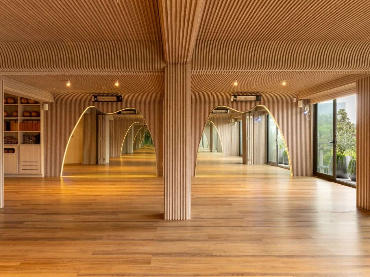 Curved light oak slatted timber ceiling and wall cladding framing illuminated arched openings in a yoga studio.