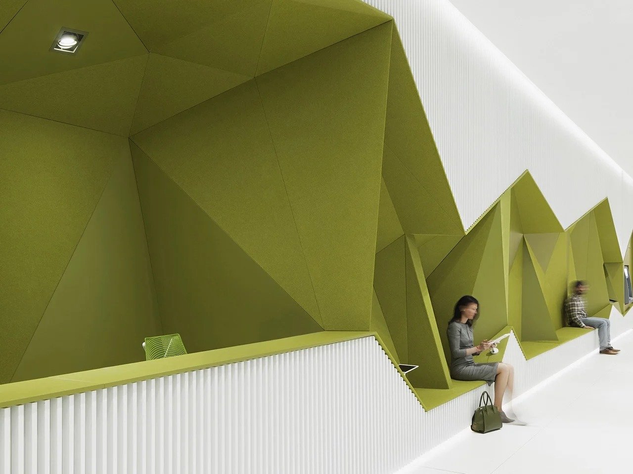 A continuous, geometric olive-green architectural ribbon wall with folded planes and integrated white ribbed seating, featuring people sitting and reading in a modern DSK Bank branch.