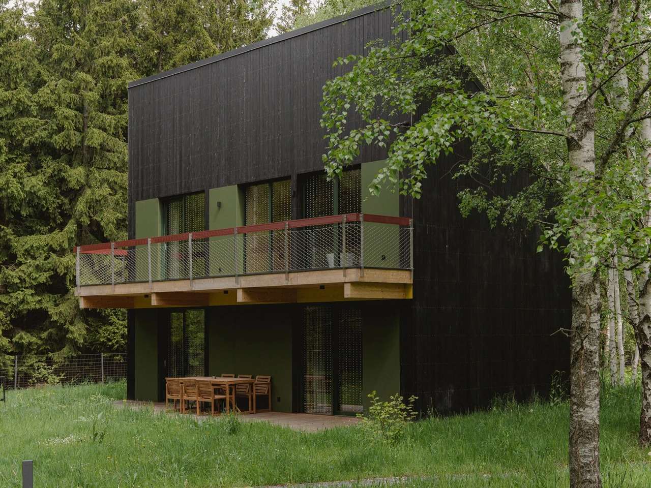Exterior view of a black wooden cabin with a second-floor balcony and wooden terrace set in a green forest landscape.