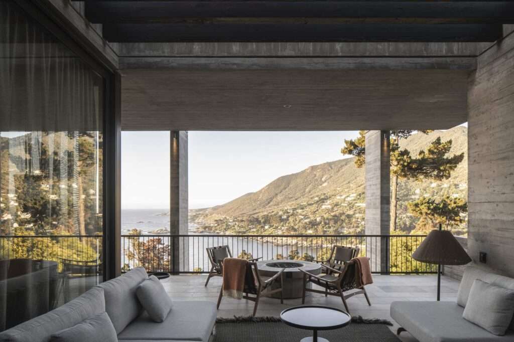 Sheltered outdoor lounge with a fire pit and seating framed by board-formed concrete columns facing coastal mountains.