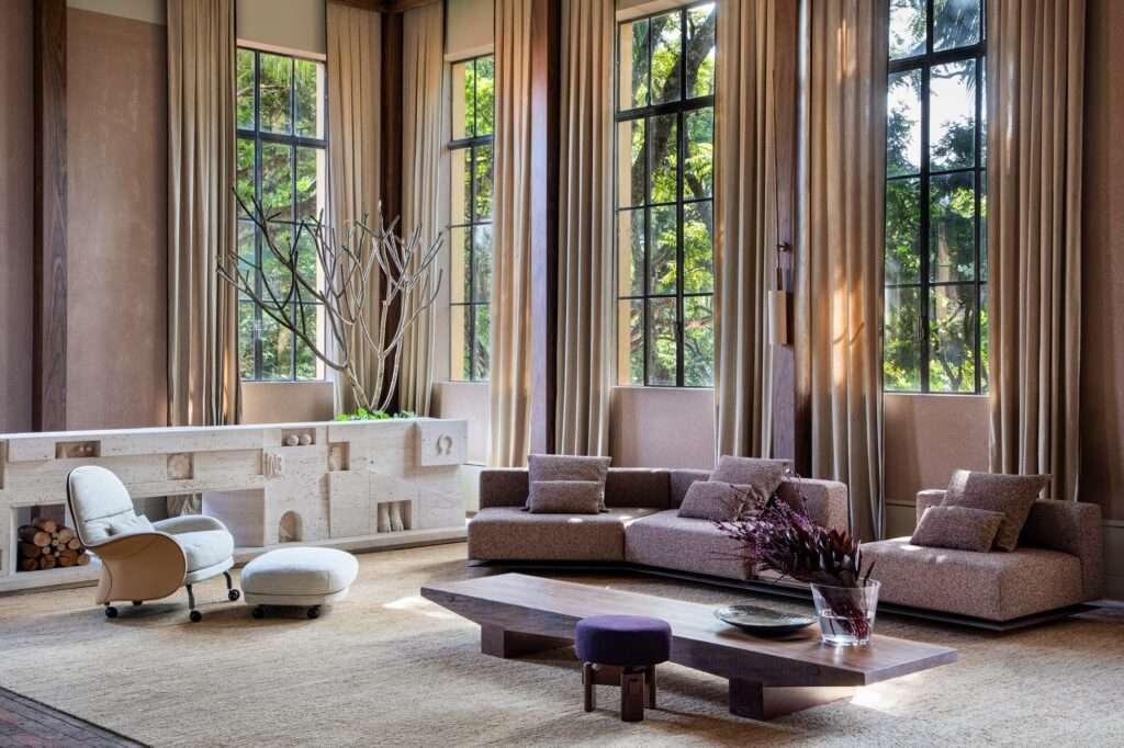 Living room with tall framed windows, a sculptural beige stone fireplace with an indoor tree, modular sofa, and low wooden coffee table.