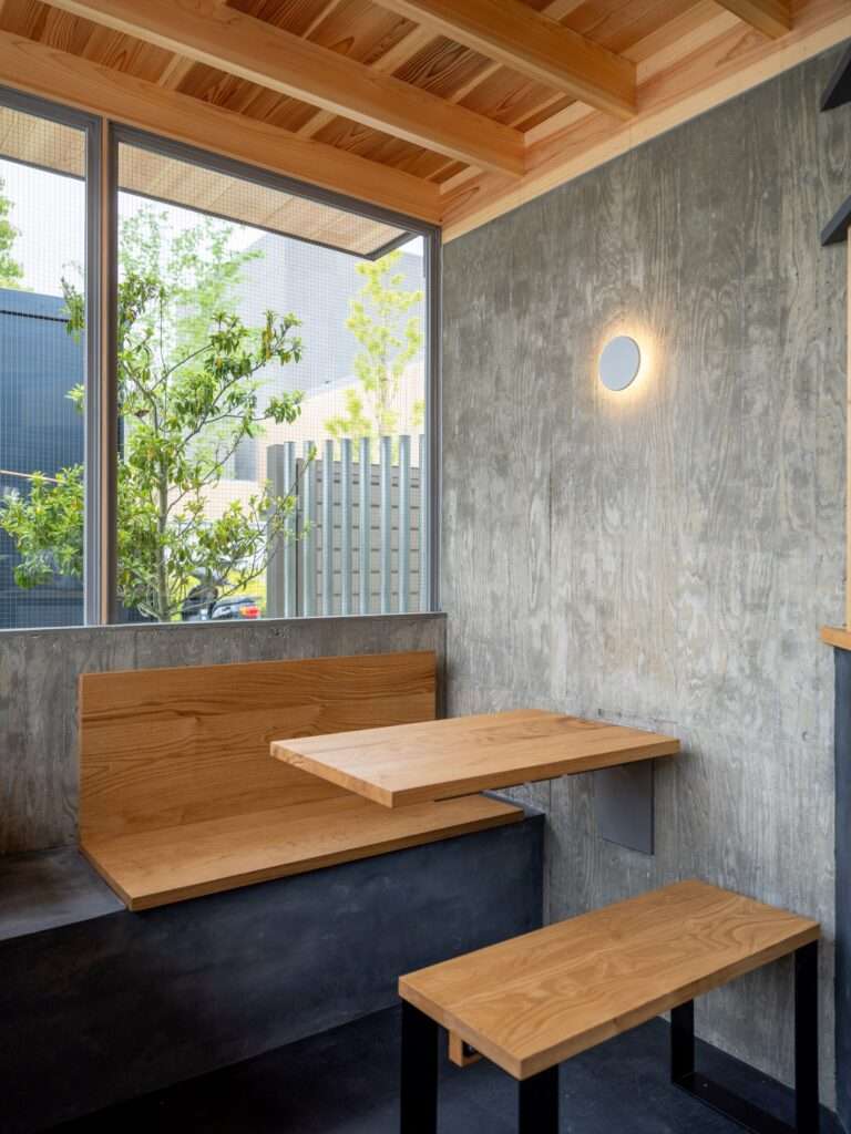 Compact wooden dining nook inside Cafe N+ with a cantilevered wooden table, matching wooden bench, board-formed concrete wall, and circular sconce light.