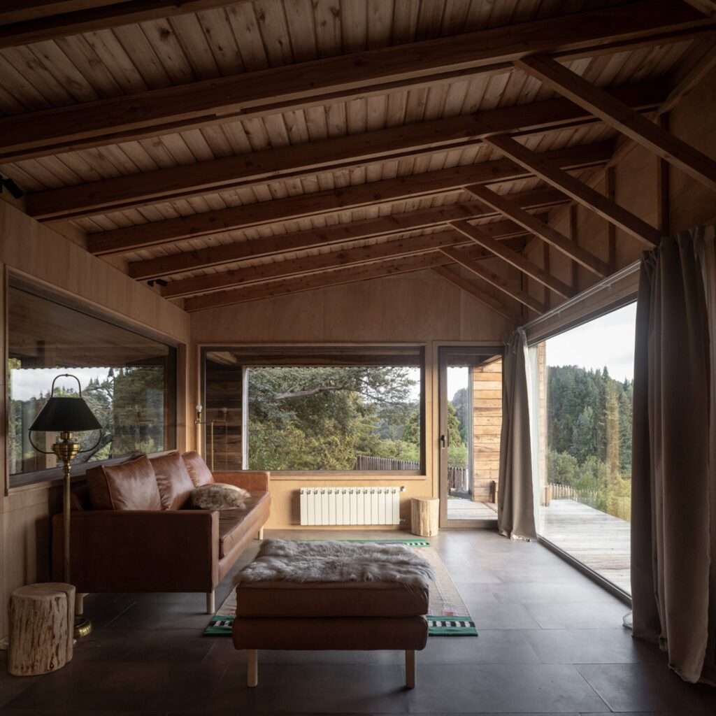 Living room interior featuring matte Guatambu wood paneling, exposed rafters, leather couch, and panoramic window facing the Patagonian forest.