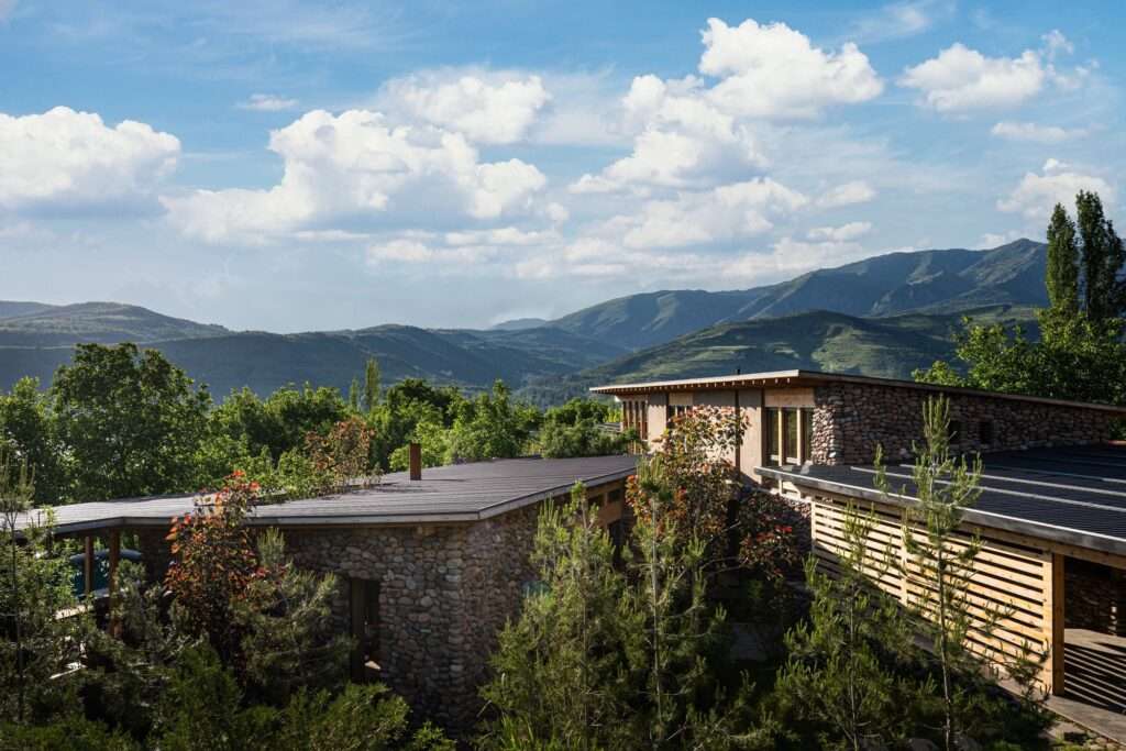 Aerial exterior view of Buladala House sloped rooflines nestled in lush greenery facing mountain landscape and Charvak Reservoir.