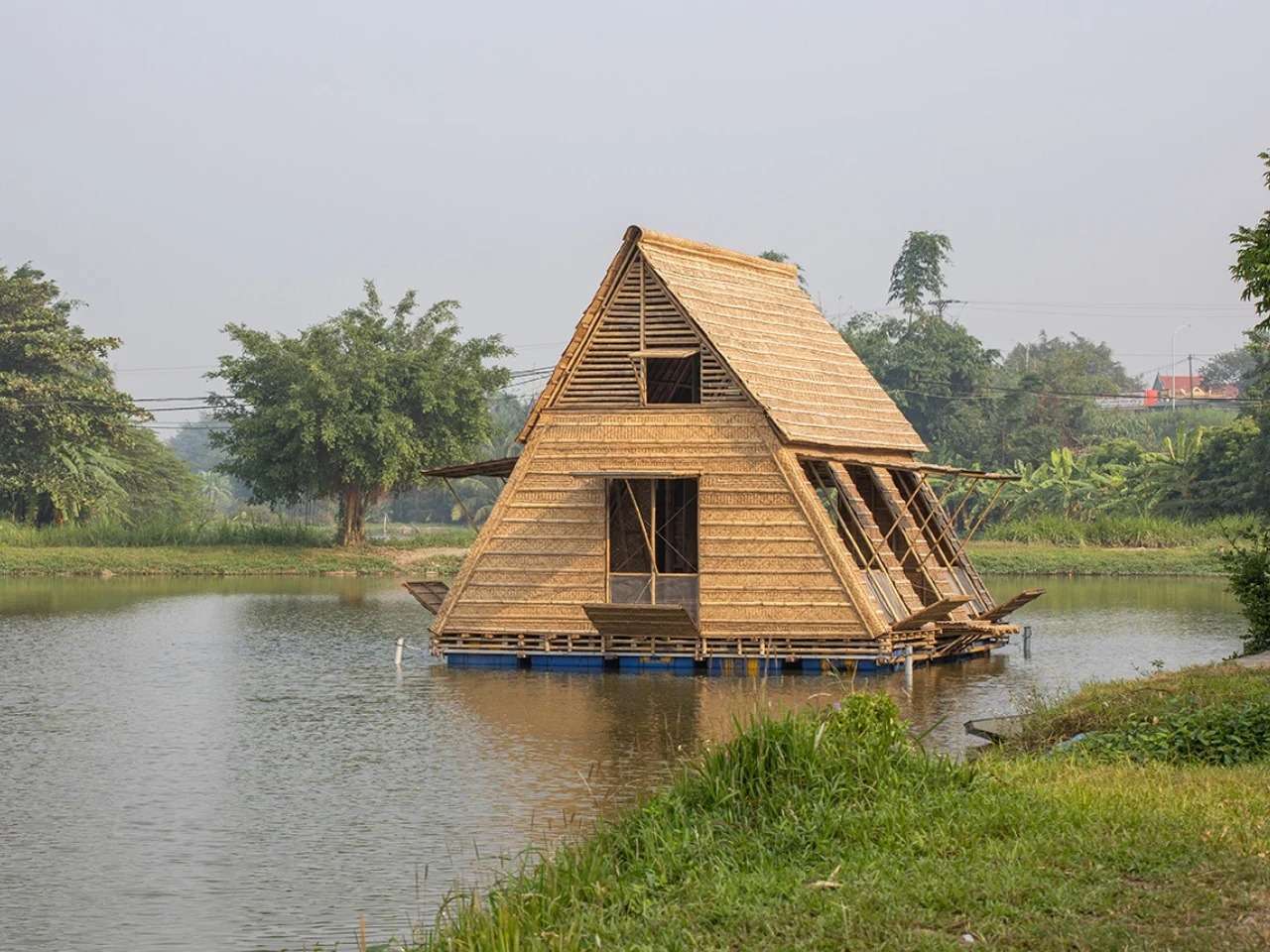 The Floating Bamboo House by H&P Architects, a triangular structure made of bamboo on a platform of blue barrels, floating on calm water in the Mekong Delta, with green banks and trees in the background.