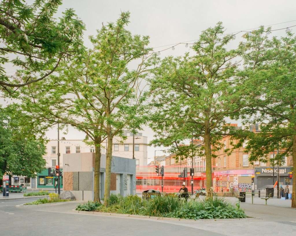 Wide view of Maida Hill civic pavilion framed by trees in a public square with red London bus motion blur in the background.