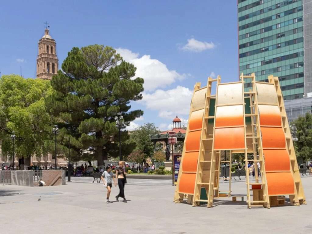 Temporary wooden arch pavilion Paso Nara installed at Plaza de Armas in Chihuahua with pedestrians walking nearby.