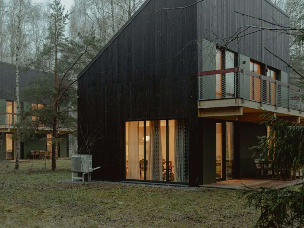 Two modern black timber cabins positioned at an angle within a forest setting during dusk with interior lights visible.