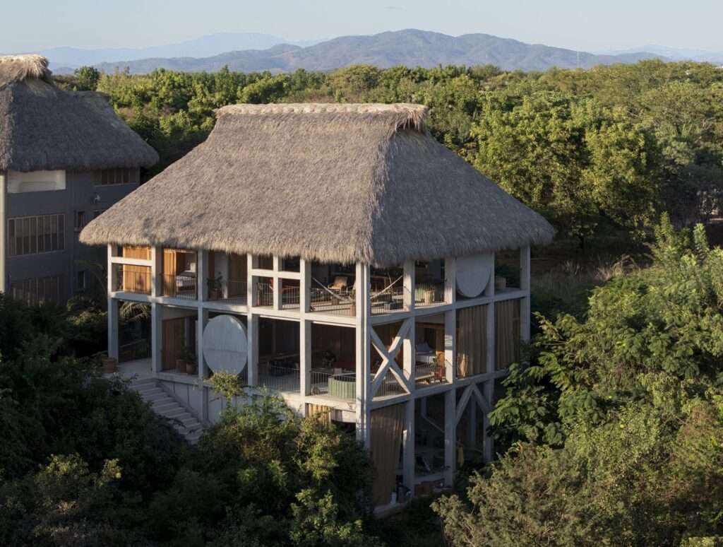 Elevated side view of Casa Eva nestled in lush green canopy with exposed concrete columns, diagonal bracing, and thatched palapa roof against distant mountains.