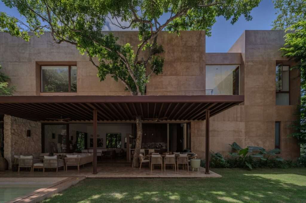 Rear elevation of Casa Sudzal showing earth-toned plaster facade, shaded terrace, swimming pool, and preserved mature tree penetrating the roof overhang.