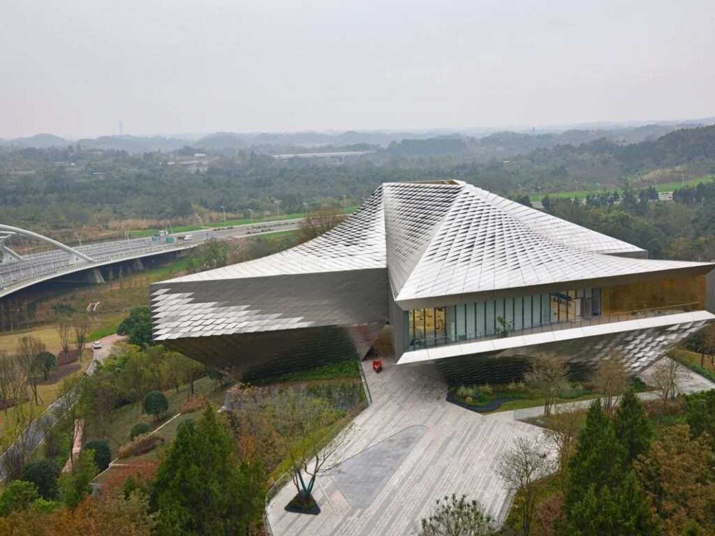 Aerial view of Chengdu Pavilion showing its three triangular wings clad in reflective stainless steel tiles set amidst a lush landscape.